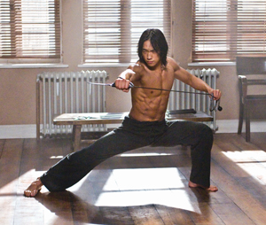 A shirtless man with long hair and toned muscles strikes a wide martial arts pose indoors, holding a sword in both hands. Sunlight streams through blinds onto the wooden floor.