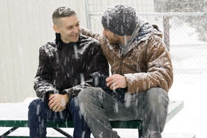 Two men sit close together on a snow-covered bench outside, one with his arm around the other. Snow falls heavily as they look at each other and smile warmly. One man is holding a black thermos.