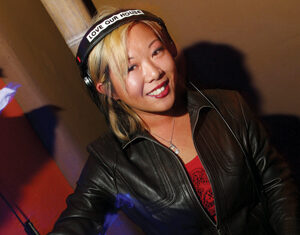 A woman wearing headphones and a black leather jacket stands at a DJ booth, smiling while mixing music on a turntable. The headphones have a label that says LOVE OUR HOUSE.