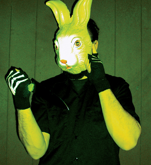 A person wearing a yellow rabbit mask and skeleton gloves holds a knife, standing against a plain wooden wall. The lighting is greenish-yellow, creating an eerie and unsettling atmosphere.