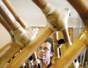 A person wearing glasses examines a structure made of bamboo poles joined together with tape. The scene appears to be indoors, possibly in a workshop, with tools and materials visible in the background.