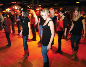 A group of people, mostly women in jeans and boots, are line dancing on a wooden floor in a dimly lit room with a crowd watching in the background.