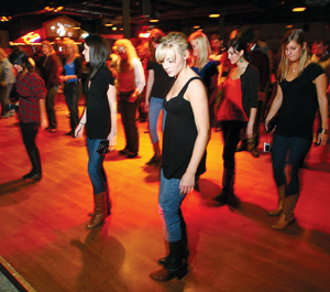 A group of people, mostly women in jeans and boots, are line dancing on a wooden floor in a dimly lit room with a crowd watching in the background.