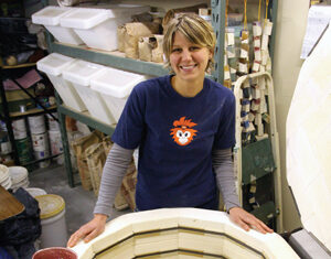 A woman smiles while standing behind an open pottery kiln filled with several red-glazed ceramic bowls and dishes. Shelves with pottery supplies and materials are visible in the background.