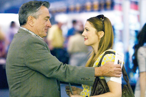 An older man in a suit warmly embraces a younger woman with long hair and a yellow-patterned top; they are smiling and making eye contact in a public indoor setting.