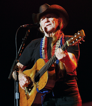 An older man wearing a black cowboy hat and black shirt plays an acoustic guitar and sings into a microphone on stage, with stage lights shining behind him.