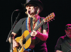 An older man with long gray hair, wearing a black cowboy hat and t-shirt, plays an acoustic guitar and sings into a microphone on stage. Another musician is partially visible in the background.