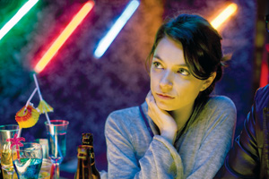 A young woman sits at a table in a dimly lit bar with colorful neon lights in the background, resting her chin on her hand and looking thoughtfully to the side. Drinks with cocktail umbrellas are on the table.