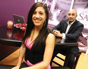 A young woman with long dark hair smiles while sitting on a bench in the foreground. A man in a suit sits at a table behind her in an office with purple walls. A laptop and a candle are on the table.