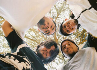 Four young men stand close together in a circle, looking down at the camera with trees and sky in the background. They appear relaxed and are dressed in casual clothes.