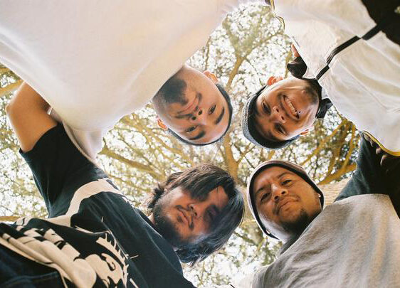Four young men stand close together in a circle, looking down at the camera with trees and sky in the background. They appear relaxed and are dressed in casual clothes.