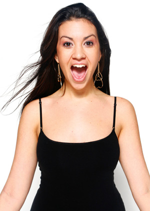 A woman with long brown hair wearing a black sleeveless top and large earrings stands against a white background, looking surprised with her mouth open and eyes wide.