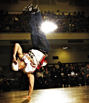 A breakdancer balances on one hand, legs extended upward, performing a dynamic move on a gym floor in front of an audience. Bright light shines behind the dancer, highlighting the action.