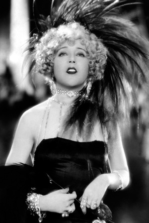 A glamorous woman in a strapless dress poses with dramatic curly hair, adorned with a large feathered headpiece, pearl necklaces, and rings, evoking classic 1920s Hollywood glamour in a black-and-white photo.
