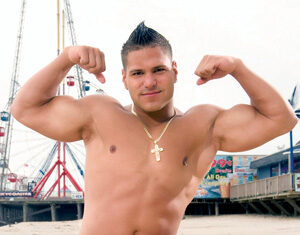 A man kneels on the sand at a beach, flexing his biceps and smiling. He is shirtless, wearing red shorts with designs and a gold chain. A pier and amusement park rides are visible in the background.