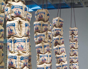 Several tall, vertical stacks of burlap sacks labeled Castillo hang from the ceiling on thick ropes in a modern art gallery space with concrete floors.