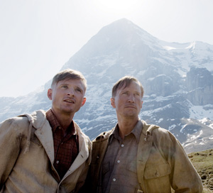 Two men wearing jackets stand outdoors with a snowy mountain peak in the background, looking off into the distance under bright sunlight.