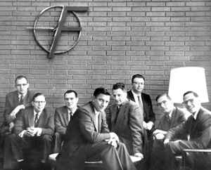 A black and white photo showing eight men in suits sitting and standing together in front of a brick wall, with a large circular logo and a floor lamp behind them.