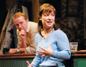 A woman in a blue sweater and plaid skirt sits on a barstool, gesturing as she speaks. A man in a beige sweater leans on the bar behind her, listening. There is a glass with a drink on the bar near the woman.