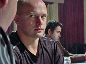 A bald man in a dark shirt looks intently at someone off-camera while sitting at a table; another man in the background is focused on something in front of him.