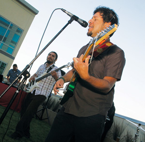 A man sings into a microphone and plays guitar while another man plays bass beside him during an outdoor performance in front of a modern building.