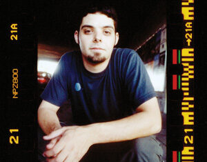 A young man with short dark hair and a goatee sits crouched, wearing a navy t-shirt and shorts. The image has a film strip border and appears to be taken in a dimly lit indoor setting.