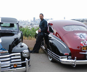 A man stands between a black vintage car and a red vintage car, with a city skyline blurred in the background. He is facing the camera, resting one hand on the red car.