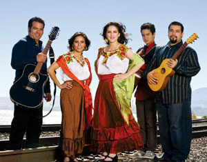 Five people stand on railroad tracks, three wearing colorful traditional Mexican clothing, two holding guitars. Mountains and a blue sky are visible in the background. They are smiling and facing the camera.