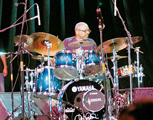 A drummer plays a blue Yamaha drum set on stage, surrounded by cymbals and microphones, with dark curtains in the background.