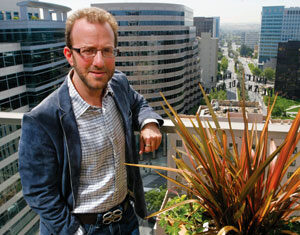A man wearing glasses, a blue jacket, and a checkered shirt stands on a rooftop balcony overlooking a cityscape with tall buildings and a street below. He is beside a planter with tall, spiky plants.