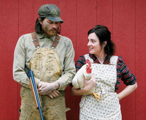 A man in overalls holding a shotgun stands next to a woman in an apron holding a white chicken. They are in front of a red wooden wall, smiling at each other.