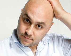 A bald man wearing a light blue button-up shirt looks at the camera while touching the top of his head with one hand, against a plain light background.