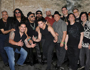 A group of thirteen men pose together in front of a textured wall, some standing and some crouching, smiling at the camera. They are dressed in casual and black clothing, with a few wearing hats and sunglasses.
