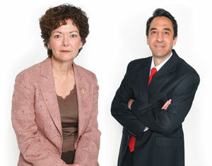 Two professionally dressed adults pose against a white background; a woman in a pink blazer sits, while a man in a dark suit and red tie stands with his arms crossed.