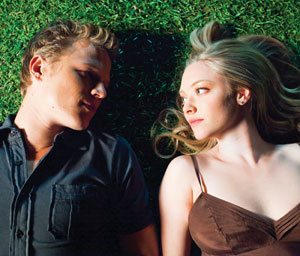 A young man and woman lie side by side on green grass, facing each other. The man wears a dark shirt, and the woman has long, blonde hair and a brown camisole. They gaze into each others eyes with soft expressions.