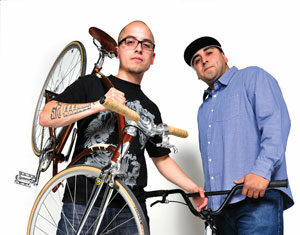 Two men pose confidently; one holds a bike on his shoulder, the other stands with his hands on the handlebars of a second bike. Both wear casual clothes and glasses against a plain white background.