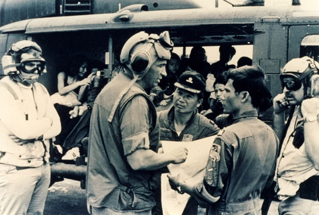 A group of military personnel gather and discuss plans next to a helicopter. Some wear helmets and headsets, while others hold papers and maps. People sit inside the helicopter in the background. The scene appears tense and focused.