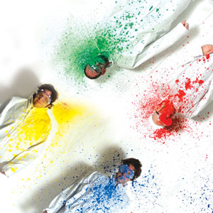 Four people in white suits lie on a white surface; each has brightly colored paint splattered over their face and chest—yellow, green, red, and blue—creating a vivid, artistic effect.
