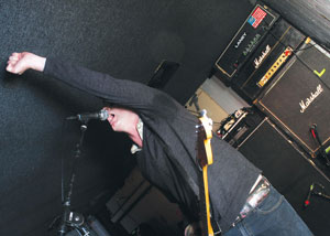 A musician performing energetically on stage, holding a guitar and raising one arm while singing into a microphone. Several large Marshall amplifiers are stacked in the background.