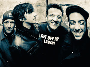 Four men stand close together, smiling and posing for the camera. One wears a shirt that says GET OFF MY LAWN! The image has a gritty, high-contrast, black-and-white filter.