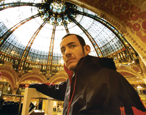 A man in a black jacket stands indoors under a large, ornate glass dome ceiling with intricate ironwork and golden decorations, looking to the side.