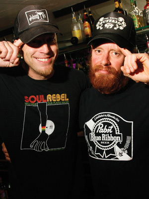 Two men in black T-shirts and caps stand side by side in a bar, smiling and touching the brims of their hats. Bottles are visible on shelves behind them. One shirt reads Soul Rebel and the other has a Pabst Blue Ribbon logo.