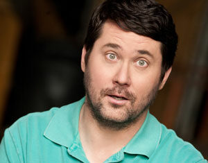 A man with short dark hair and facial hair wears a light green polo shirt, standing with his arms crossed and an expressive, surprised look on his face against a dark, blurred background.