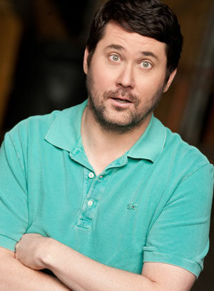 A man with short dark hair and facial hair wears a light green polo shirt, standing with his arms crossed and an expressive, surprised look on his face against a dark, blurred background.