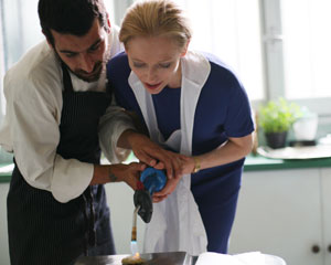 A man in a chef’s uniform helps a woman in a blue dress use a kitchen torch, both leaning over a counter in a brightly lit kitchen.