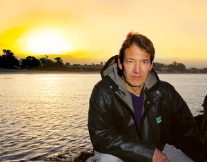 A man in a dark jacket kneels by the water’s edge at sunset, with the glowing sky and distant trees reflecting on the calm surface behind him.