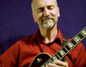 A man with a short gray beard and mustache, wearing a red shirt, plays an electric guitar with his eyes closed and a content expression, in front of a dark purple background.