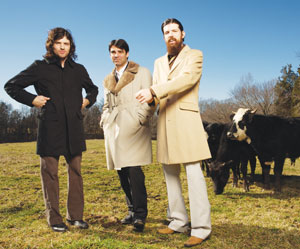 Three men in long coats stand in a grassy field with several black cows behind them. The sky is clear and blue, and trees are visible in the background.
