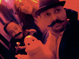 Three people with fake mustaches pose for a selfie in red and purple lighting; one holds a plush toy. The mood appears playful and festive.