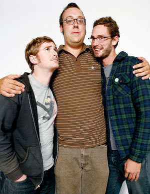 Three men stand together against a plain white background. The man in the middle wears glasses and a striped polo shirt, while the two men on either side hug him and smile. They all appear to be friends and look casual.